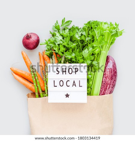 Fresh organic vegetables in eco craft paper shopping bag with text Shop Local on lightbox flat lay, top view with copy space on gray background. Sustainable lifestyle. Zero waste plastic free concept.