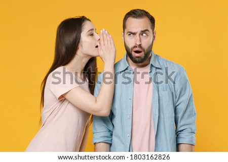 Perplexed young couple friends man woman in pastel blue casual clothes whispering secret behind hand on ears, sharing news isolated on yellow background, studio portrait.