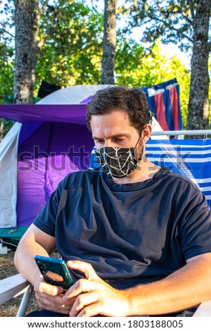 Photo of a young and attractive man wearing a face mask and checking his phone while sitting on a camping chair. Relax, holidays