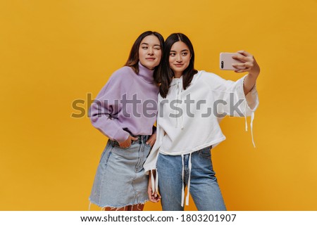 Asian girl in denim skirt and purple sweater winks. Attractive brunette woman in jeans and white hoodie holds phone. Friends take selfie on orange background.