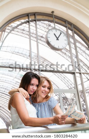 Two young beautiful smiling women standing in a waiting room of a train station looking at train tickets and hugging each other on the background of big wall clocks at daytime
