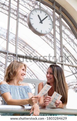 Two young beautiful smiling women standing in a waiting room of a train station with train tickets in hands and looking at each other on the background of big wall clocks at daytime. Travel concept