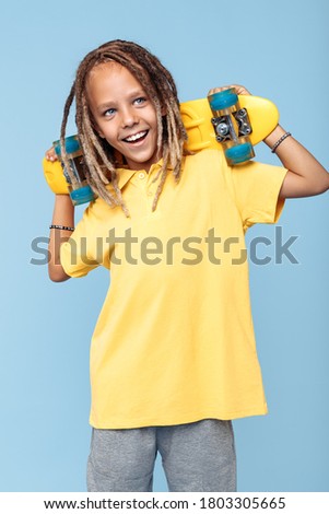 Friendly little boy with african dreads holding skateboard on shoulders and smile over blue background.