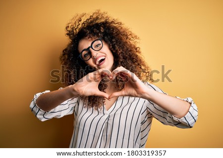 Young beautiful woman with curly hair and piercing wearing striped shirt and glasses smiling in love doing heart symbol shape with hands. Romantic concept.