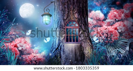 Magical fantasy elf or gnome house in tree with window and lantern, bench in enchanted fairy tale forest with fabulous fairytale blooming pink rose flower garden and shiny glowing moon rays in night Royalty-Free Stock Photo #1803335359