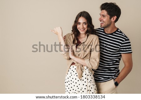 Image of cheerful beautiful couple pointing finger aside and smiling isolated over beige background