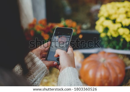 Stylish hipster woman taking photo of pumpkins and autumn flowers. Girl photographing on phone rustic halloween street decor, hands close up. Happy Thanksgiving and Halloween