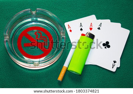 no smoking sign in a glass ashtray next to  a cigarette with a green lighter on playing cards. Top view, green backround