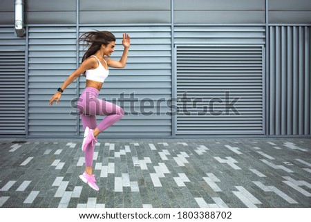 Young woman with fit body jumping and running against grey background. Female model in sportswear exercising outdoors. Modern young woman in sports clothing jumping while exercising outdoors Royalty-Free Stock Photo #1803388702
