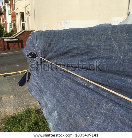 Square format photo of well worn tarps covering yellow skip with rope cord tied around the skip. Old tarpaulin covering an object with rope tied around the object on road 