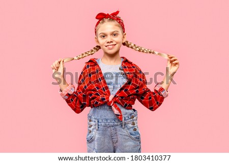 Happy child girl in casual checkered shirt standing isolated on pastel pink background with copy space, holding pigtails, smiling cheerful and looking at camera