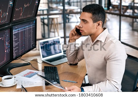 Focused businessman talking on modern smartphone, looking at monitor, checking cryptocurrency information data on finance market graph, working in office