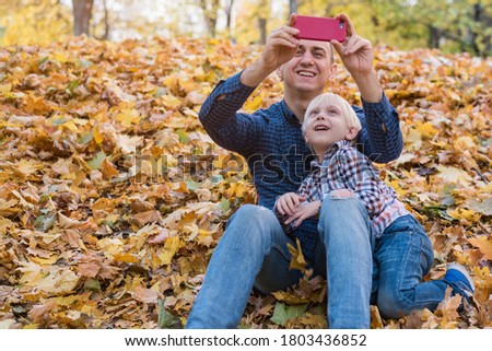 Father and son sitting on autumn leaves and make pictures on phone