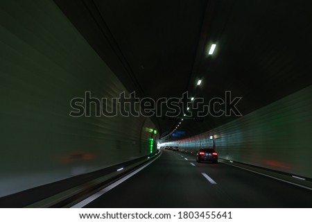 Cars on a Swiss highway tunnel, curvy road, light coming from the end of it. Mild blur on the road, focus in car. Road signs with no visual information. 