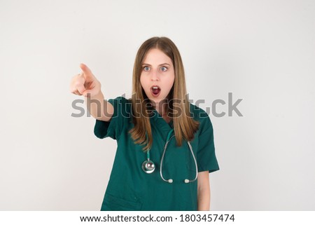 Female doctor wearing a green scrubs and stethoscope is on white background Pointing with finger surprised ahead, open mouth amazed expression, something on the front.