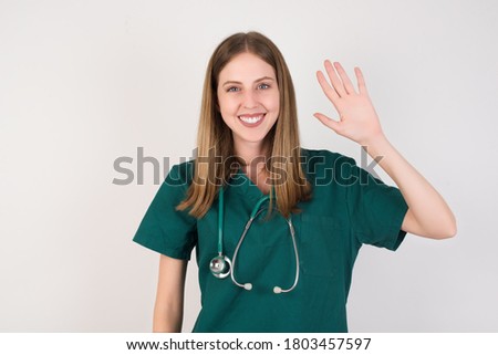 Female doctor wearing a green scrubs and stethoscope is on white background Waiving saying hello happy and smiling, friendly welcome gesture.
