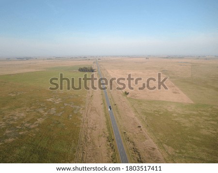 Aerial view of a two-lane road, with green fields to the sides and the blue sky in the background.