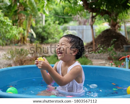 Asian cute child boy laughing while playing water in blue bowl with relaxing face and wet hair in rural nature with water splash. Young kid having happy moment in summer. Family activity at home.