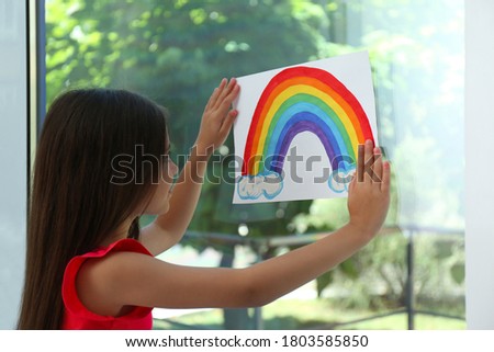 Little girl with picture of rainbow near window indoors.  Stay at home concept