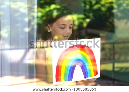 Little girl with picture of rainbow near window, view from outdoors. Stay at home concept