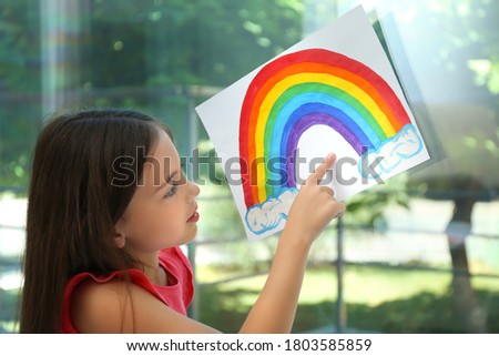 Little girl with picture of rainbow near window indoors.  Stay at home concept