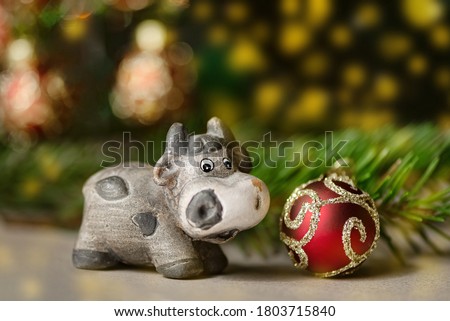 Cute ceramic bull as symbol of New Year 2021 on blurred golden garland light bokeh background next to fir branch and Christmas tree decorated with red ball