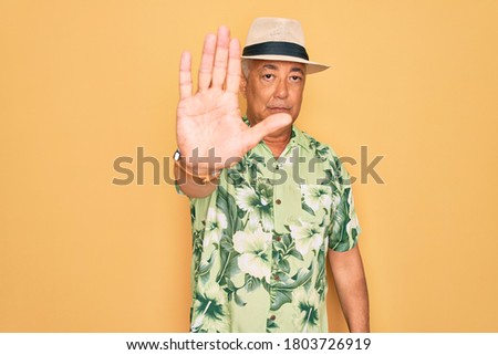 Middle age senior grey-haired man wearing summer hat and floral shirt on beach vacation doing stop sing with palm of the hand. Warning expression with negative and serious gesture on the face.