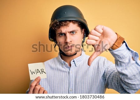Young blond businessman with curly hair wearing helmet holding paper with war message with angry face, negative sign showing dislike with thumbs down, rejection concept