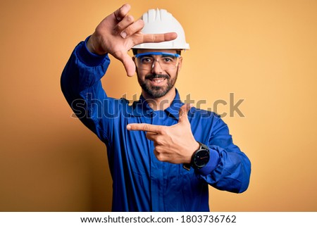 Mechanic man with beard wearing blue uniform and safety glasses over yellow background smiling making frame with hands and fingers with happy face. Creativity and photography concept.
