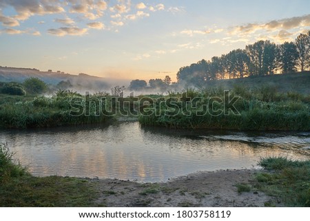 Summer time. Morning dawn over the river in a hazy, thoughtful haze. Beautiful view of the forest and river covered with fog early in the morning. The sun's rays of light.