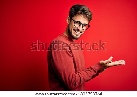 Young handsome man with beard wearing glasses and sweater standing over red background pointing aside with hands open palms showing copy space, presenting advertisement smiling excited happy
