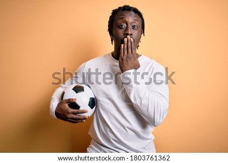 Young african american player man playing soccer holding football ball over yellow background cover mouth with hand shocked with shame for mistake, expression of fear, scared in silence, secret