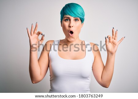 Young beautiful woman with blue fashion hair wearing casual t-shirt over white background looking surprised and shocked doing ok approval symbol with fingers. Crazy expression