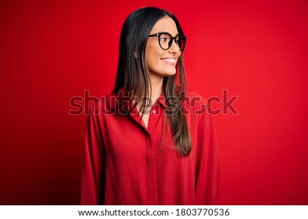 Young beautiful brunette woman wearing casual shirt and glasses over red background looking away to side with smile on face, natural expression. Laughing confident.