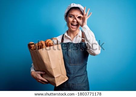 Young beautiful baker woman with blue eyes wearing apron holding paper bag with croissants with happy face smiling doing ok sign with hand on eye looking through fingers
