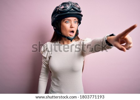 Young beautiful motorcyclist woman with blue eyes wearing moto helmet over pink background Pointing with finger surprised ahead, open mouth amazed expression, something on the front