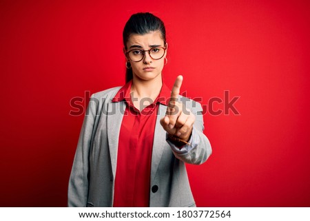 Young beautiful brunette businesswoman wearing jacket and glasses over red background Pointing with finger up and angry expression, showing no gesture