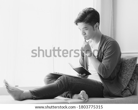 Young Asian man writing in a book when using laptop
