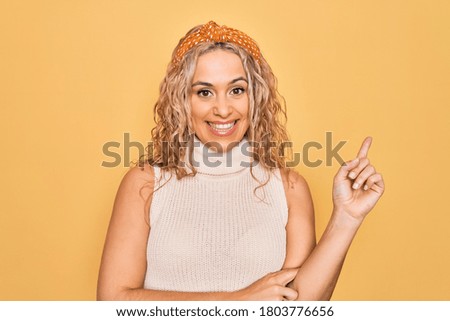 Beautiful blonde woman wearing casual t-shirt and diadem over isolated yellow background with a big smile on face, pointing with hand and finger to the side looking at the camera.