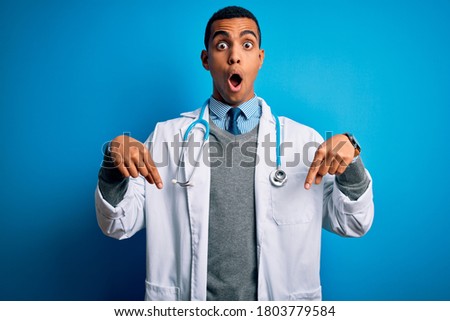 Handsome african american doctor man wearing coat and stethoscope over blue background Pointing down with fingers showing advertisement, surprised face and open mouth