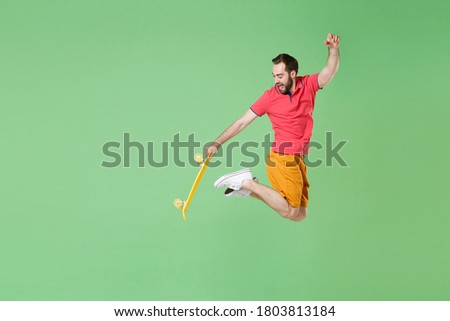 Full length of sporty strong young bearded man 20s in casual pink polo shirt jumping high in air and holding in hand yellow skateboard, looking back isolated on green background, studio portrait. 