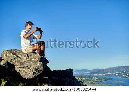 Dark teenager sitting on a rock photographing the landscape with his mobile. Young boy in a white t-shirt and light shorts watching the landscape from a rock. Happy teenager watching the landscape