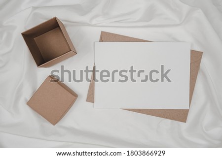 Blank white paper on brown paper envelope and carton box put on white cloth. Mock-up of horizontal blank greeting card. Top view of Craft envelope on white background. Flat lay minimalism