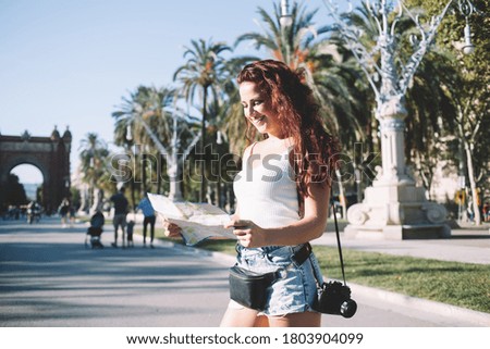 Happy redhead woman with photo camera smiling and examining map while standing on street on sunny weekend day in Barcelona City