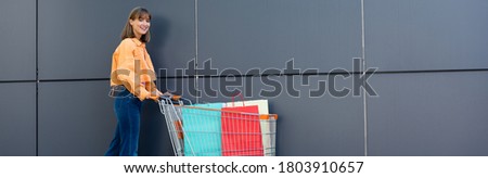 Panoramic concept of young woman looking at camera near shopping bags in cart and building