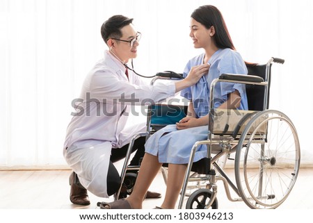 Male doctor using a stethoscope checking lungs and heart of female patient sitting on wheelchair. Doctor checking heart beat of female patient. Doctor using stethoscope checking patient's vitals sign.