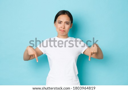 Skeptical grimacing asian woman looking displeased, pointing fingers down and smirking dissatisfied with product, showing bad advertisement promo, standing blue background