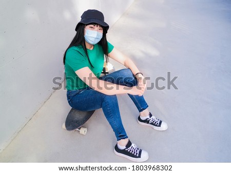Beautiful young asian woman sitting on a skateboard and wearing protective face mask - Coronavirus lifestyle and health care