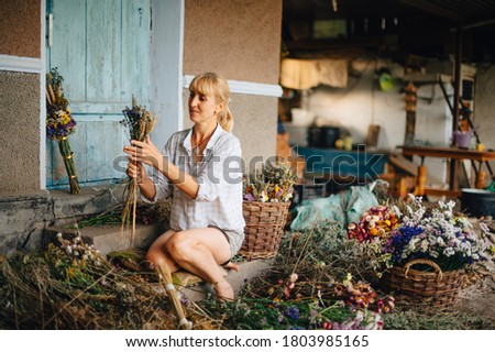 Beautiful adult woman sits in the country among the dried flowers and creates a beautiful holiday bouquet with a serious face. Silly woman makes beautiful bouquets of dried flowers.