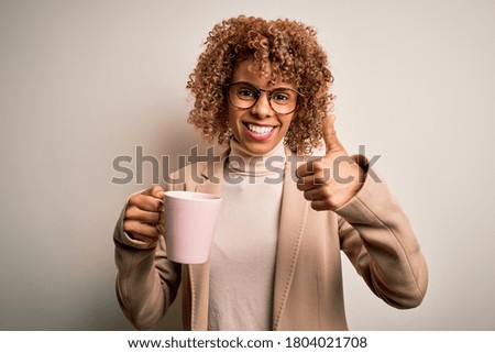 Young african american curly woman drinking mug of coffee over isolated white background happy with big smile doing ok sign, thumb up with fingers, excellent sign